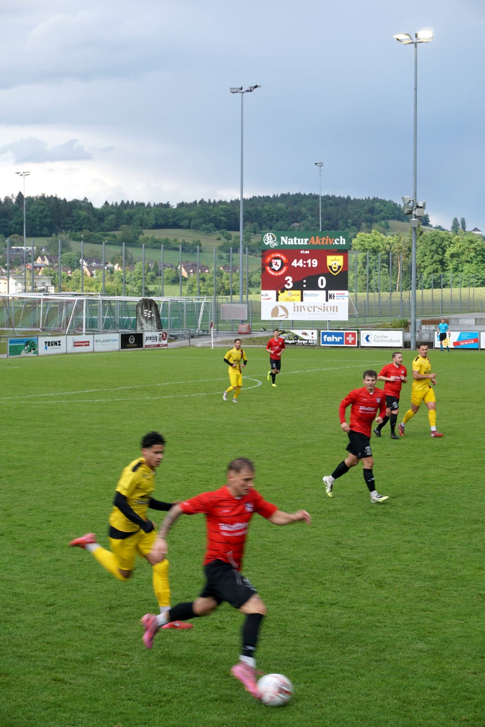 Match action on a football pitch, with the LED scoreboard showing 3:0 and the match clock in the background, sponsor ads below